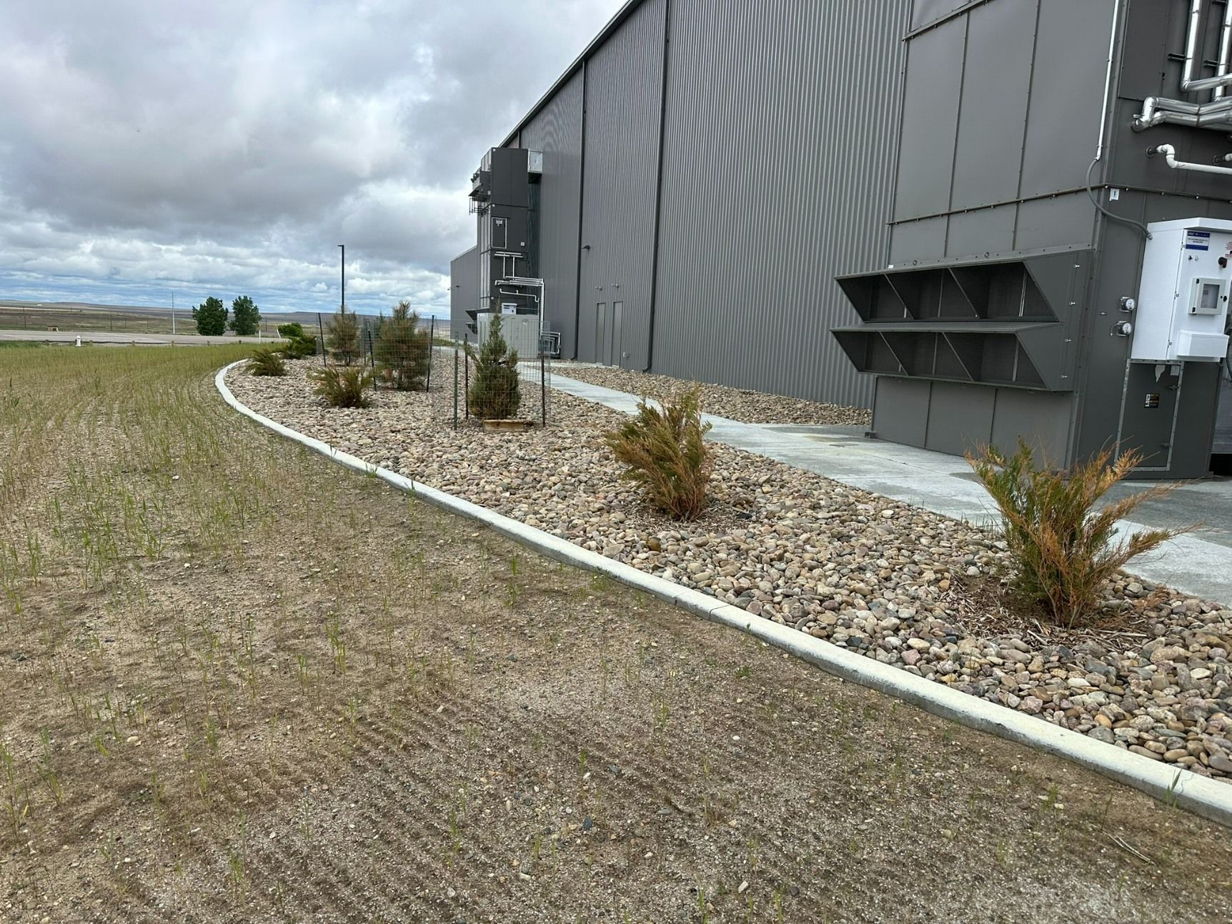 A grey industrial building with a curved concrete border containing gravel and small shrubs next to a dry grass lawn.