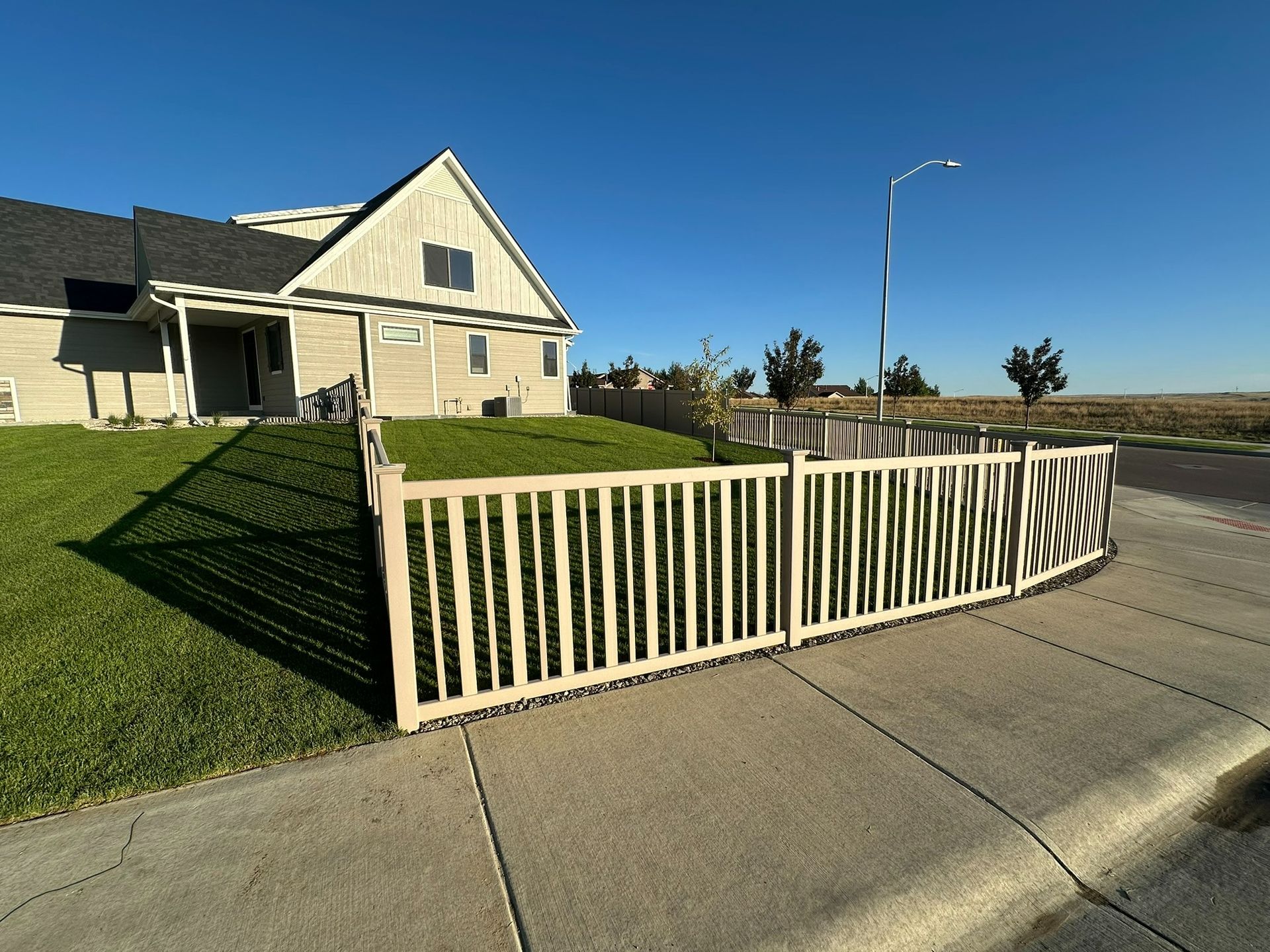 A tan picket fence stands in front of a house with a green lawn under a clear blue sky.