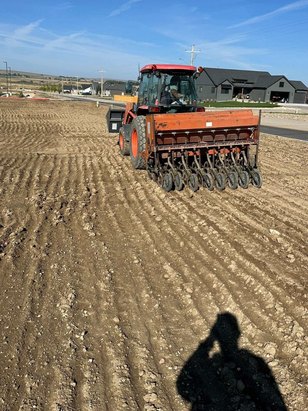 An orange tractor pulls a seed drill across a tilled field in a residential area on a sunny day.