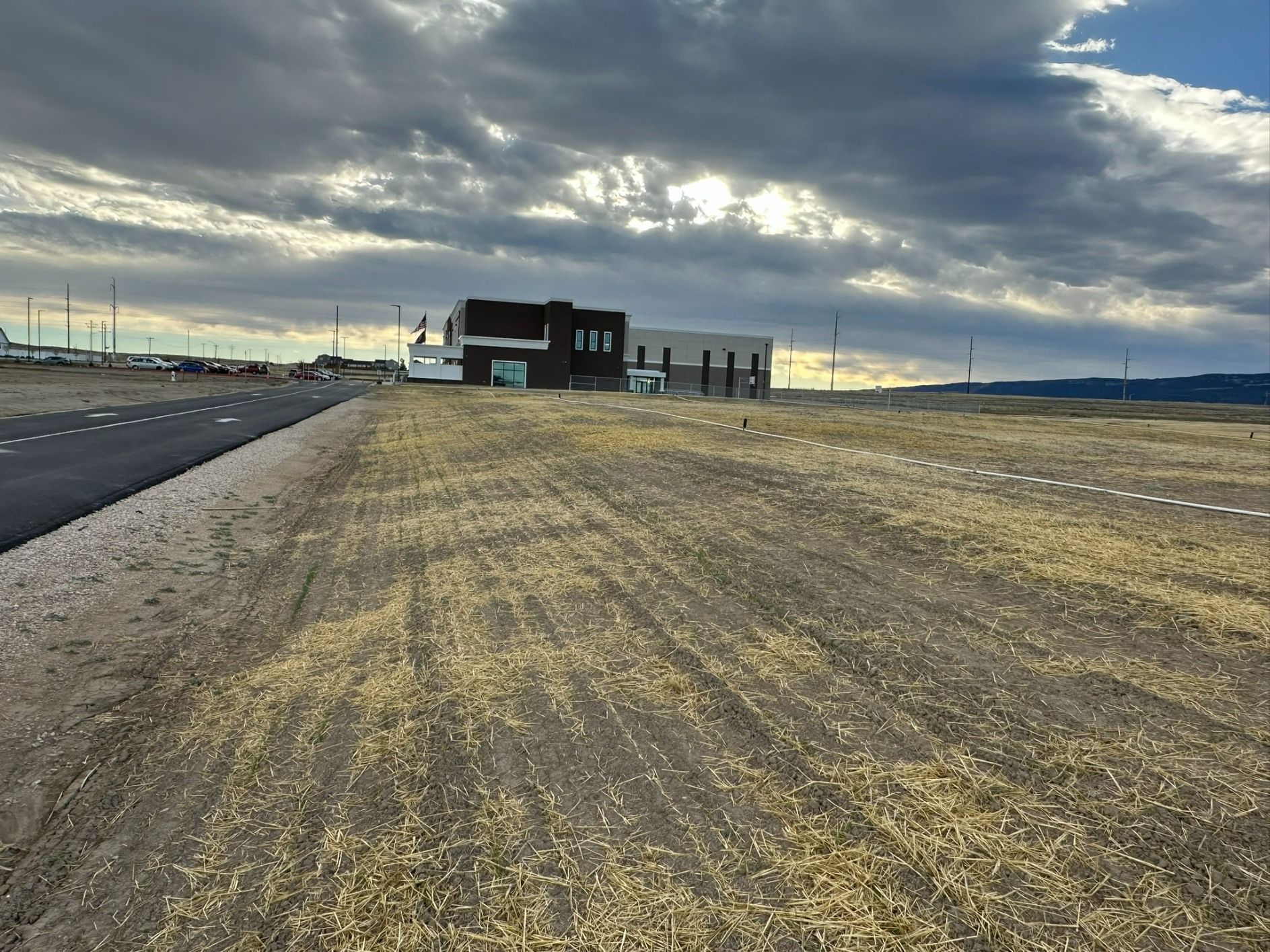A paved road runs beside a grassy field leading toward a modern, dark-sided building under a cloudy, dramatic sky.