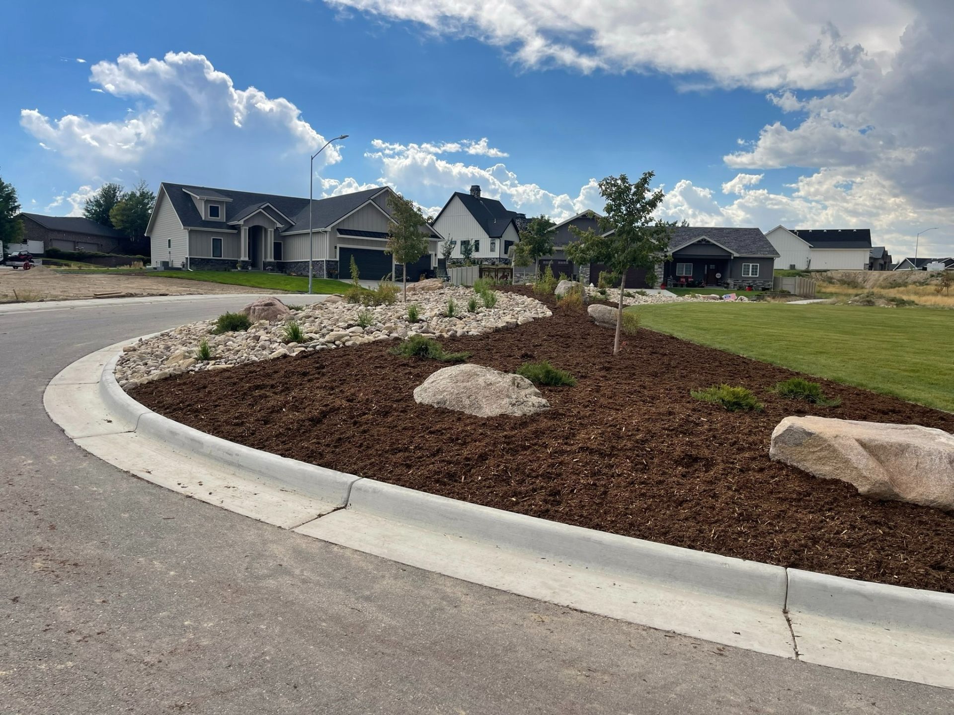 A landscaped street corner with brown mulch, large decorative rocks, and small shrubs, bordering a neighborhood road.