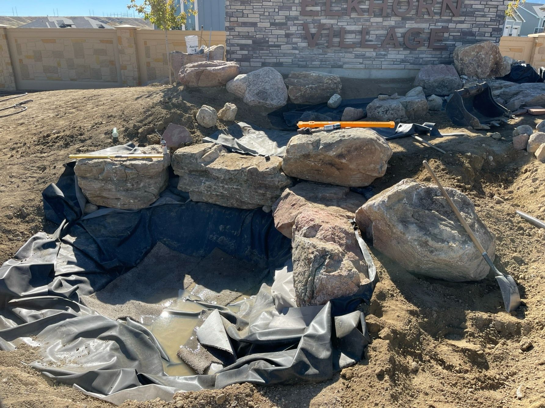 Construction of a stone pond feature with black liner in front of a stone sign wall in a dirt landscape.