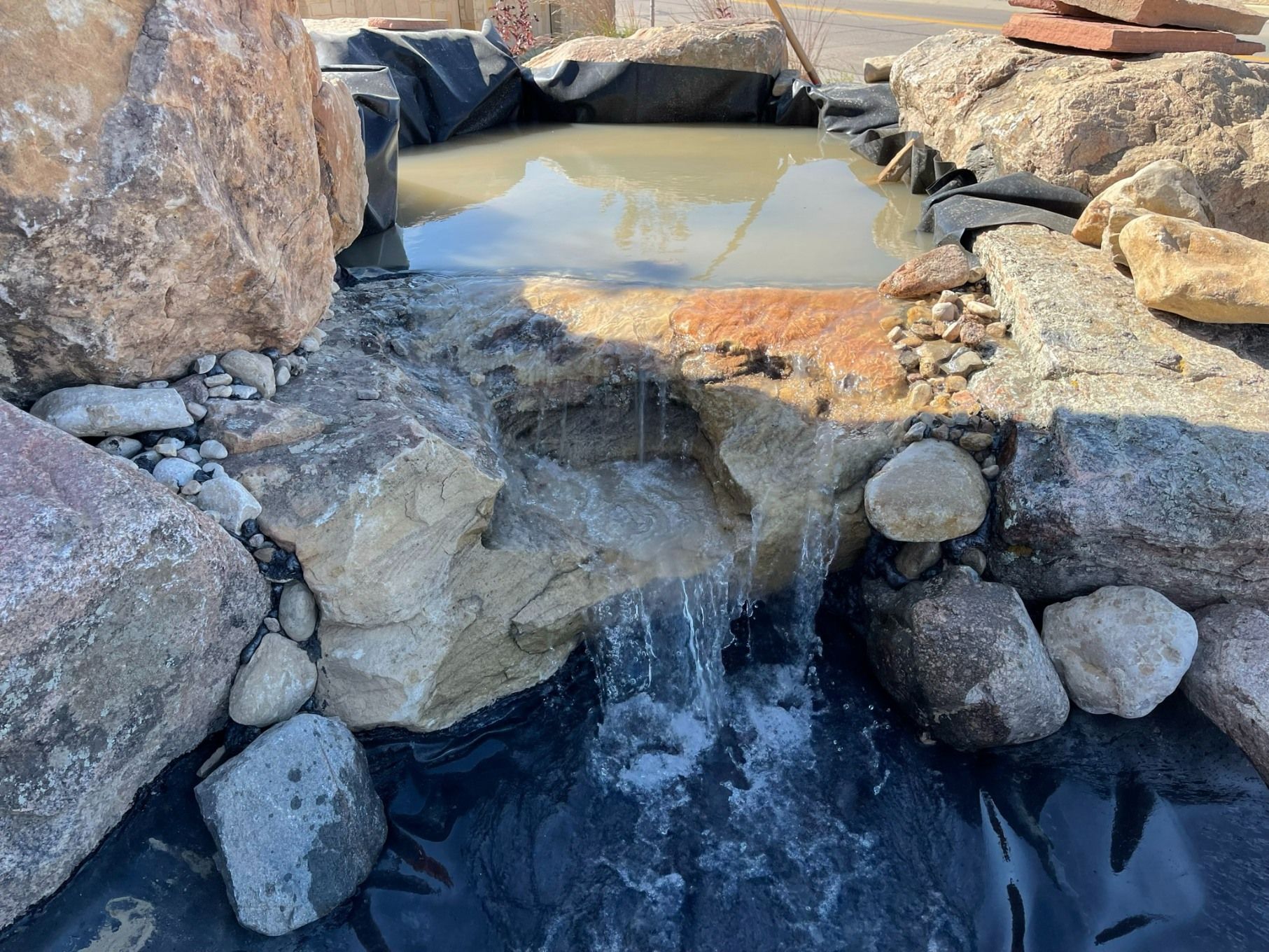 Water cascades from a rock basin into a pond lined with black landscape fabric in an outdoor setting.