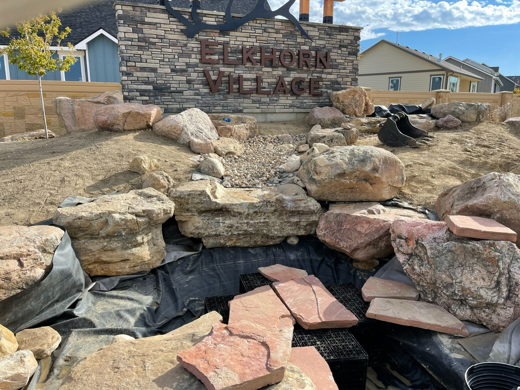An Elkhorn Village stone sign sits above a decorative rock fountain and pool currently under construction.