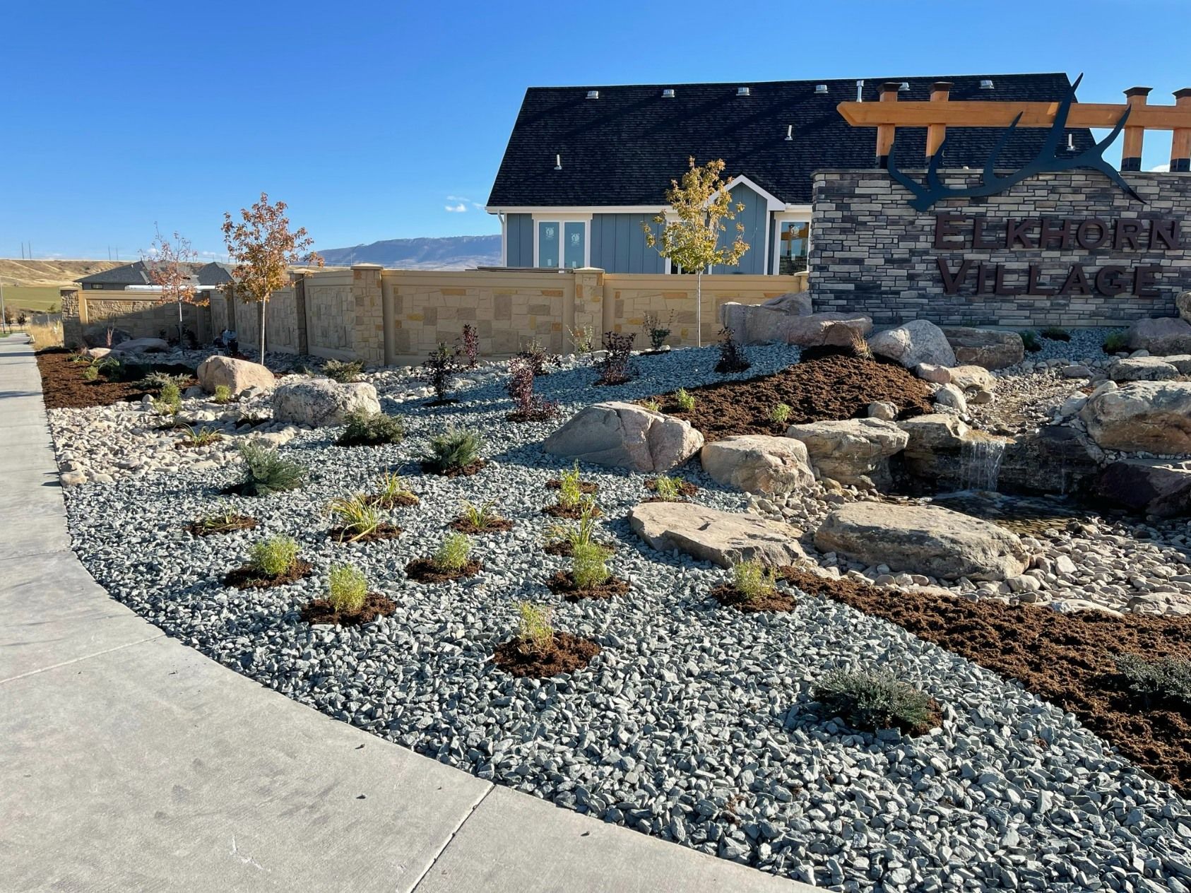 A landscaping feature with gray gravel, small shrubs, and rocks, next to a concrete sidewalk and a stone neighborhood sign.