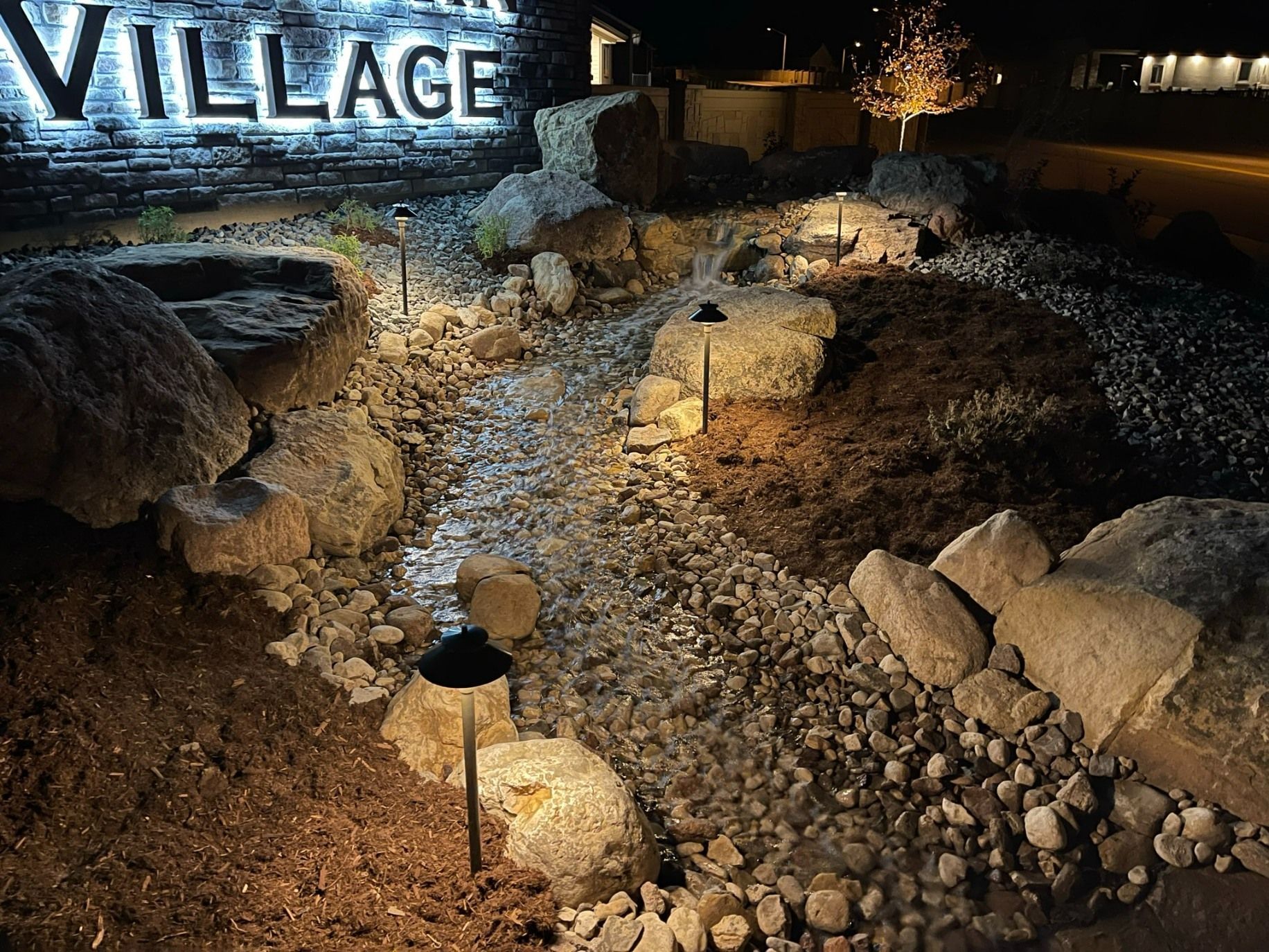 Nighttime view of a stone-lined dry creek bed illuminated by small pathway lights below a sign reading 
