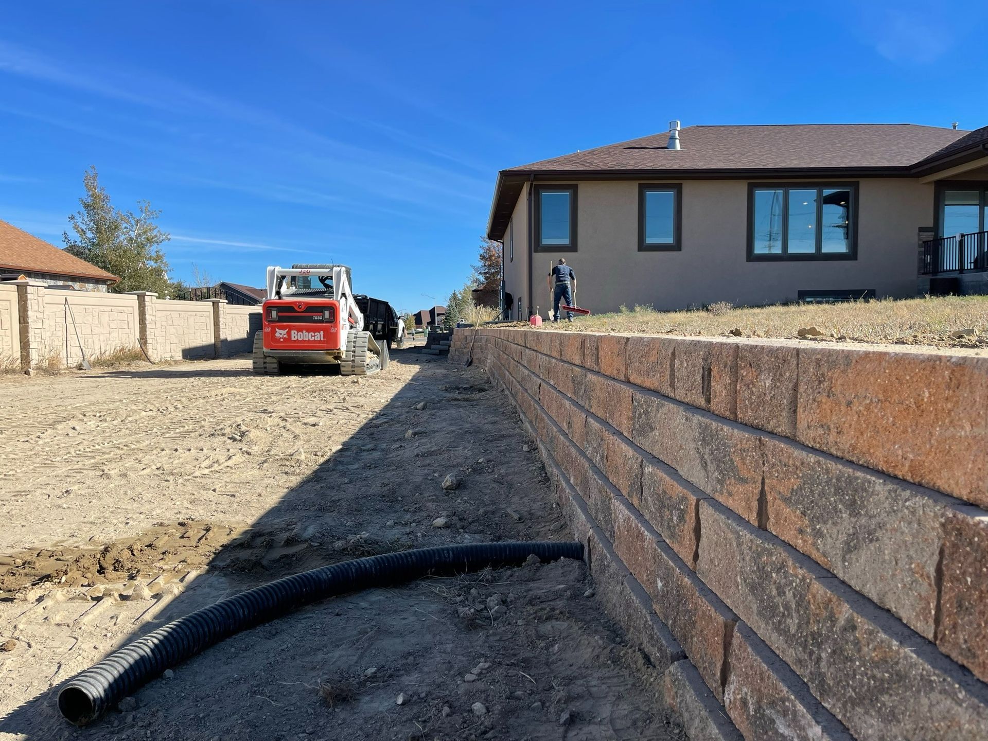 A construction site featuring a newly built retaining wall alongside a house and a small skid steer loader.