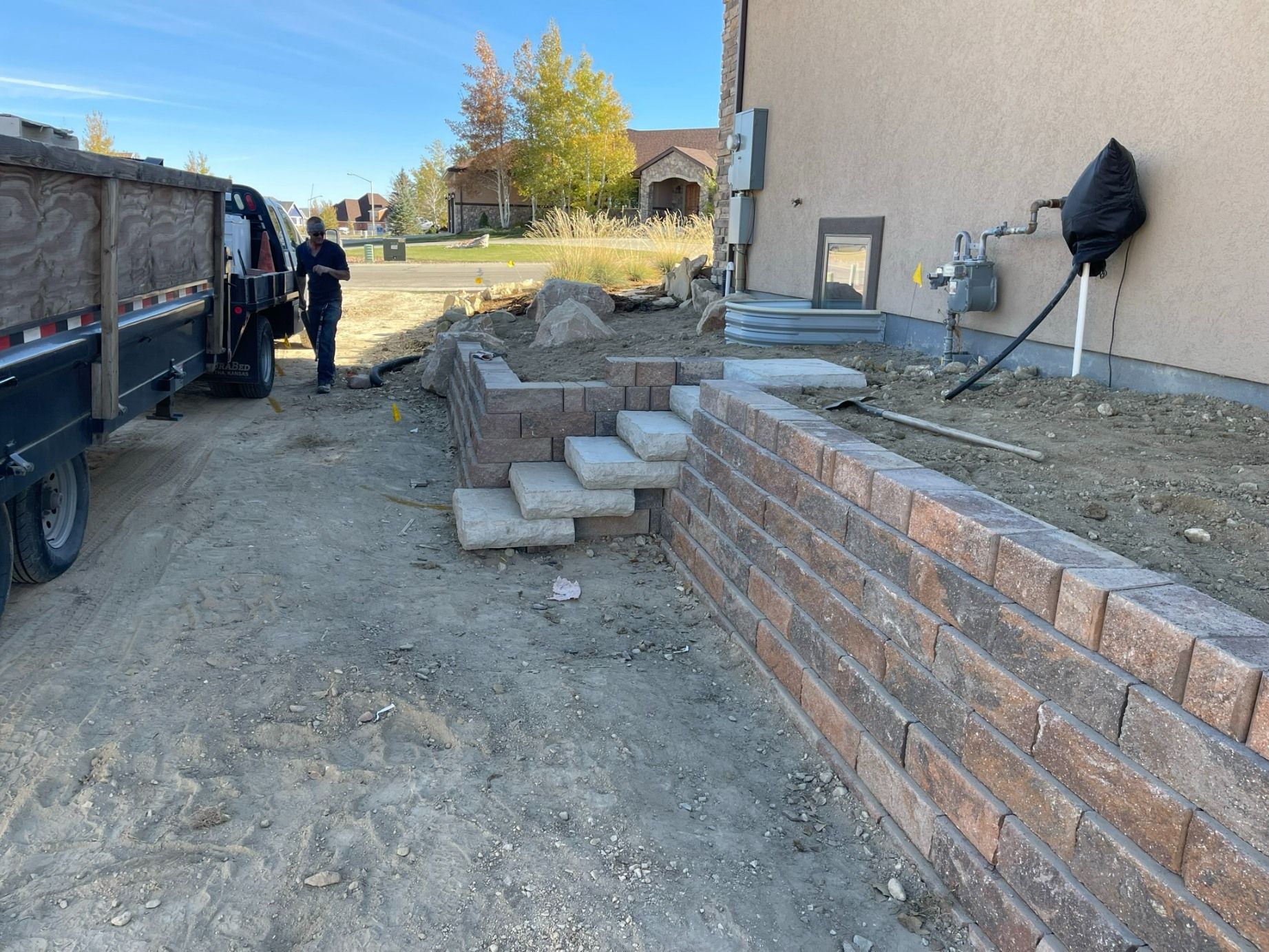 A worker stands next to a truck beside a newly constructed stone retaining wall and staircase outside a suburban home.