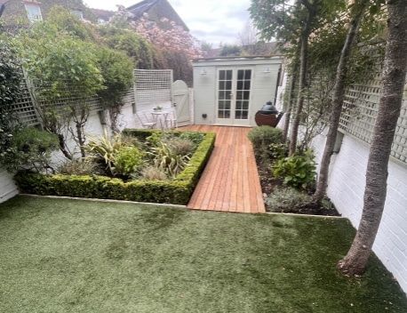 Small backyard garden with artificial turf, a wooden path, and a white shed surrounded by plants