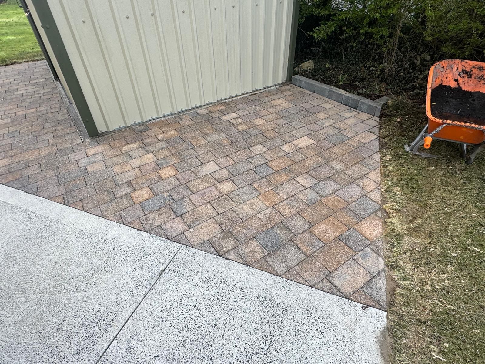 Paver walkway beside a shed, meeting a concrete slab and grassy yard, with an orange wheelbarrow at right