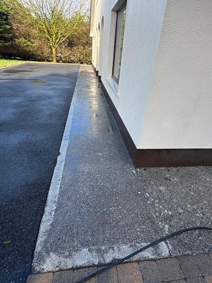 Narrow concrete walkway beside a white building, with a curb and paved driveway leading to trees.