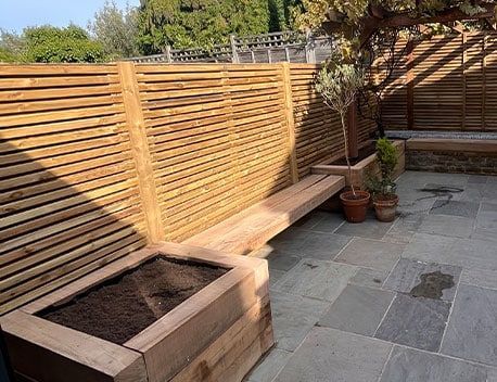 Sunny patio with wooden slat fencing, raised planter box, potted plants, and stone paving.
