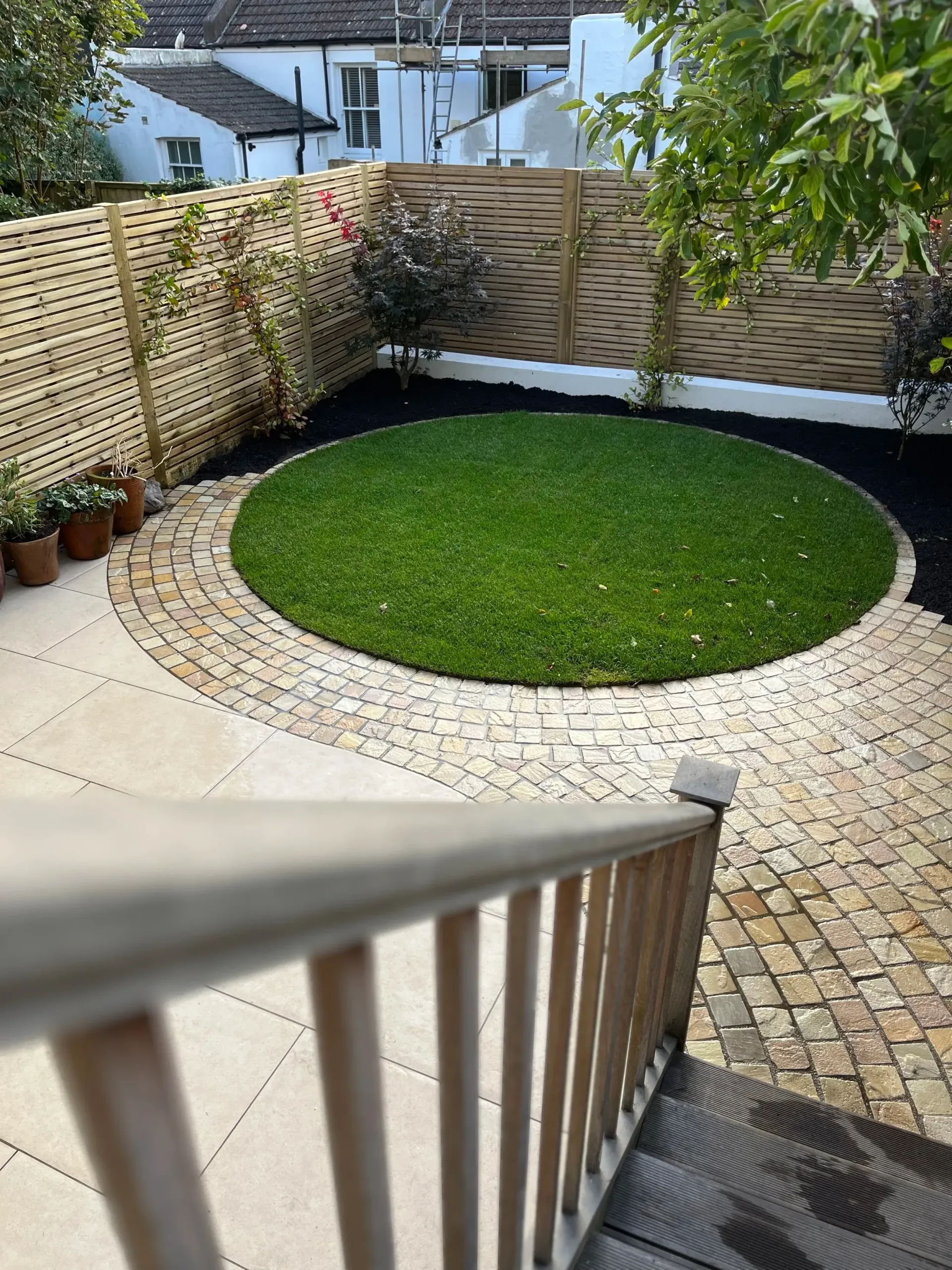 Backyard patio with circular lawn, curved stone border, wooden fence, and benches under a tree.