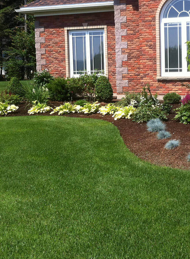 Brick house with white-trimmed windows, green lawn, and landscaped flower beds along the front yard.
