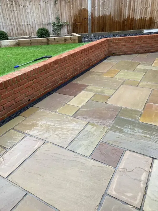 Stone patio with tan pavers beside a low red brick retaining wall and grassy yard.