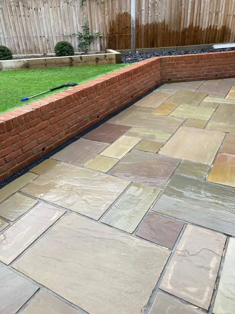 Backyard patio with beige stone pavers and a red brick retaining wall beside a green lawn.