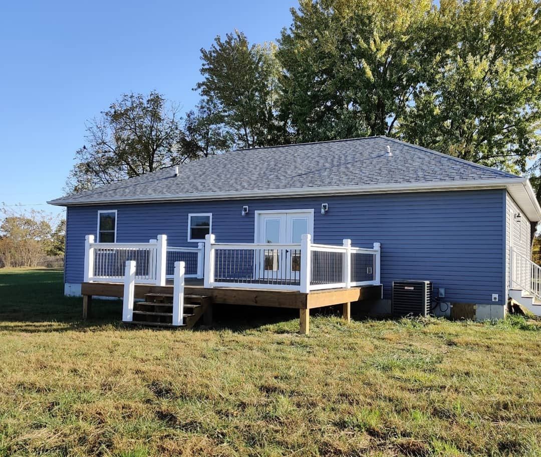A blue house with a white deck is sitting in the middle of a grassy field.