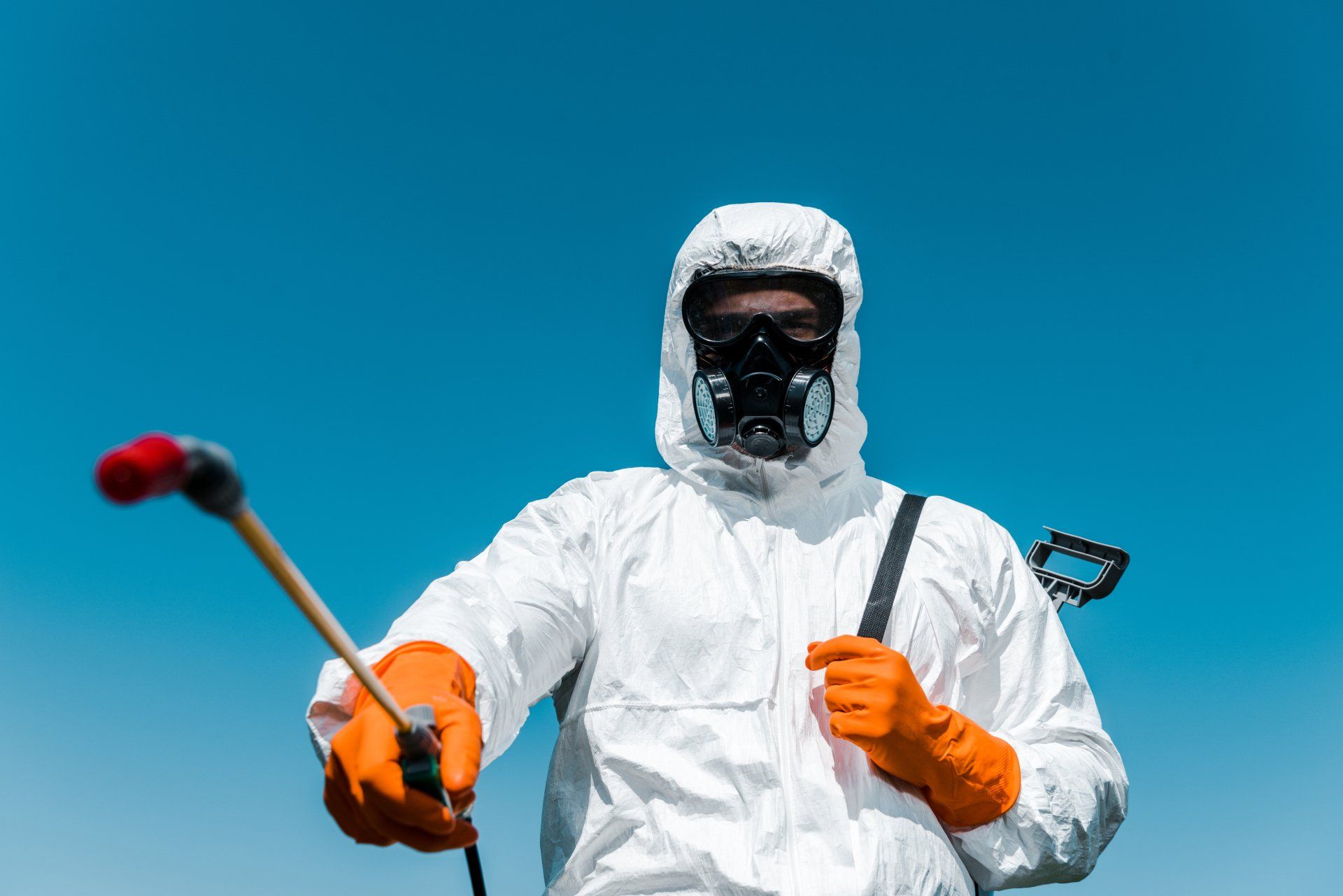 Une personne en combinaison de protection et respirateur pulvérisant avec une buse, sur fond de ciel bleu.