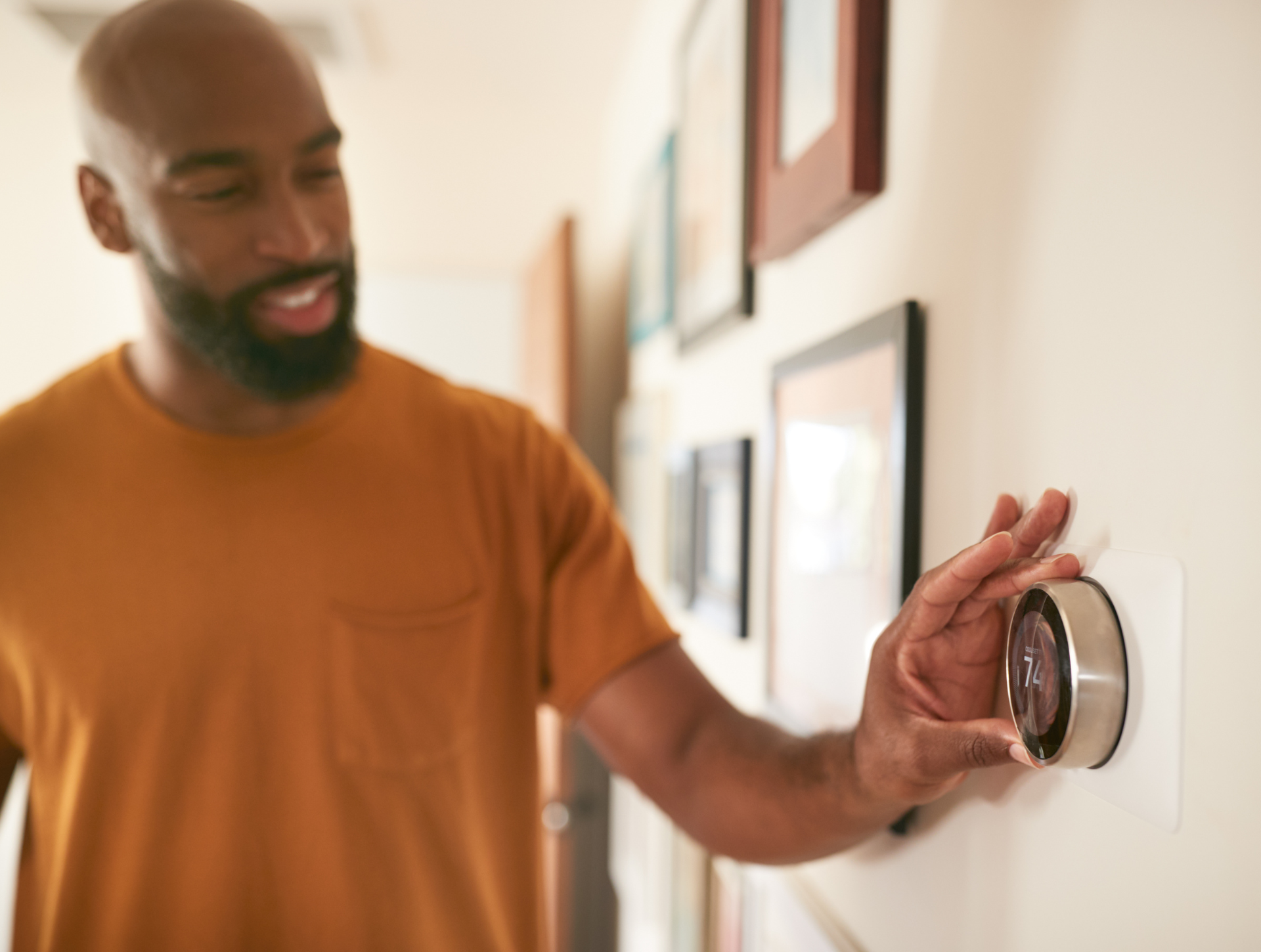A man in an orange shirt adjusts a smart thermostat on a beige wall.