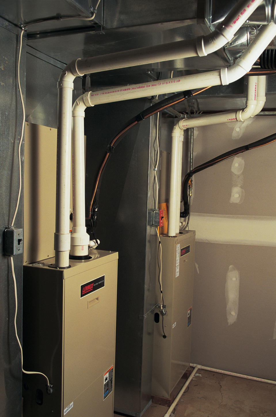 Two beige furnaces with white PVC pipes in a basement.