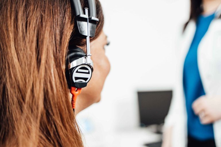 Woman wearing headset, facing away, in office setting. Another person in background.