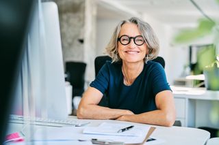 Woman with short gray hair and glasses smiles at desk in office.
