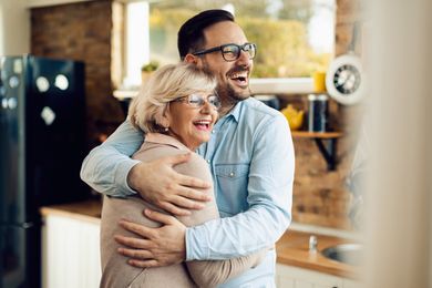 Man hugging and laughing with a woman in a kitchen; they both wear glasses and smile.