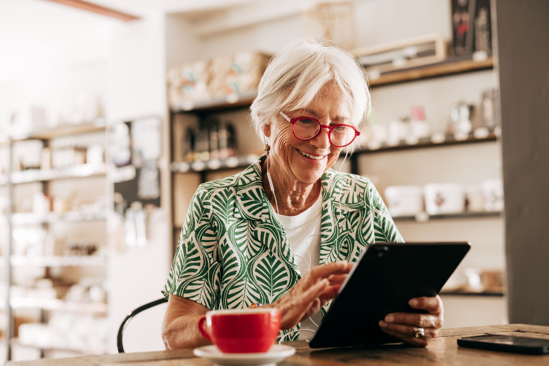 Woman smiling while using a tablet at a table in a cafe, with a red coffee cup.