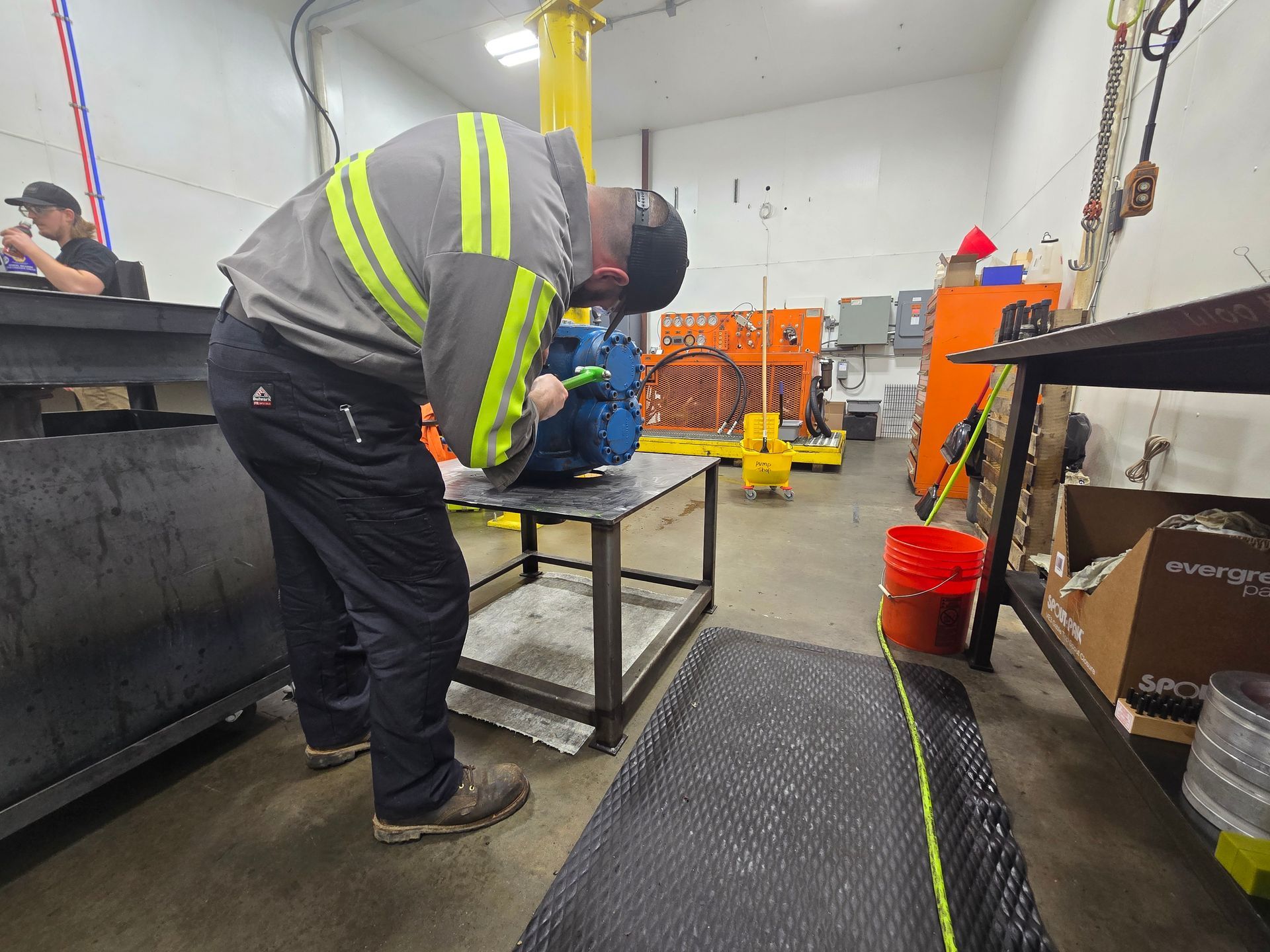 Worker in reflective vest bends over a table in a workshop with tools and equipment.