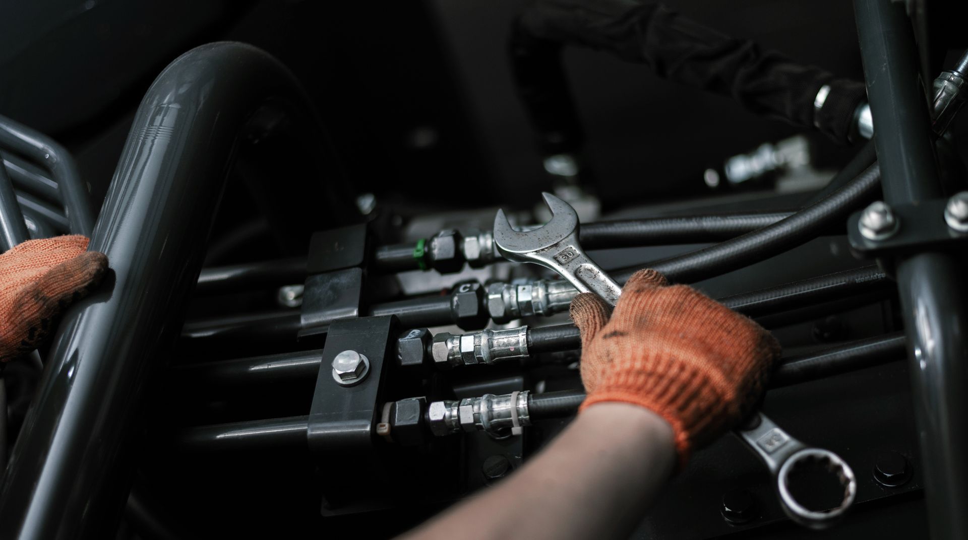 Hands in orange gloves tighten a fitting on black tubes with a wrench.