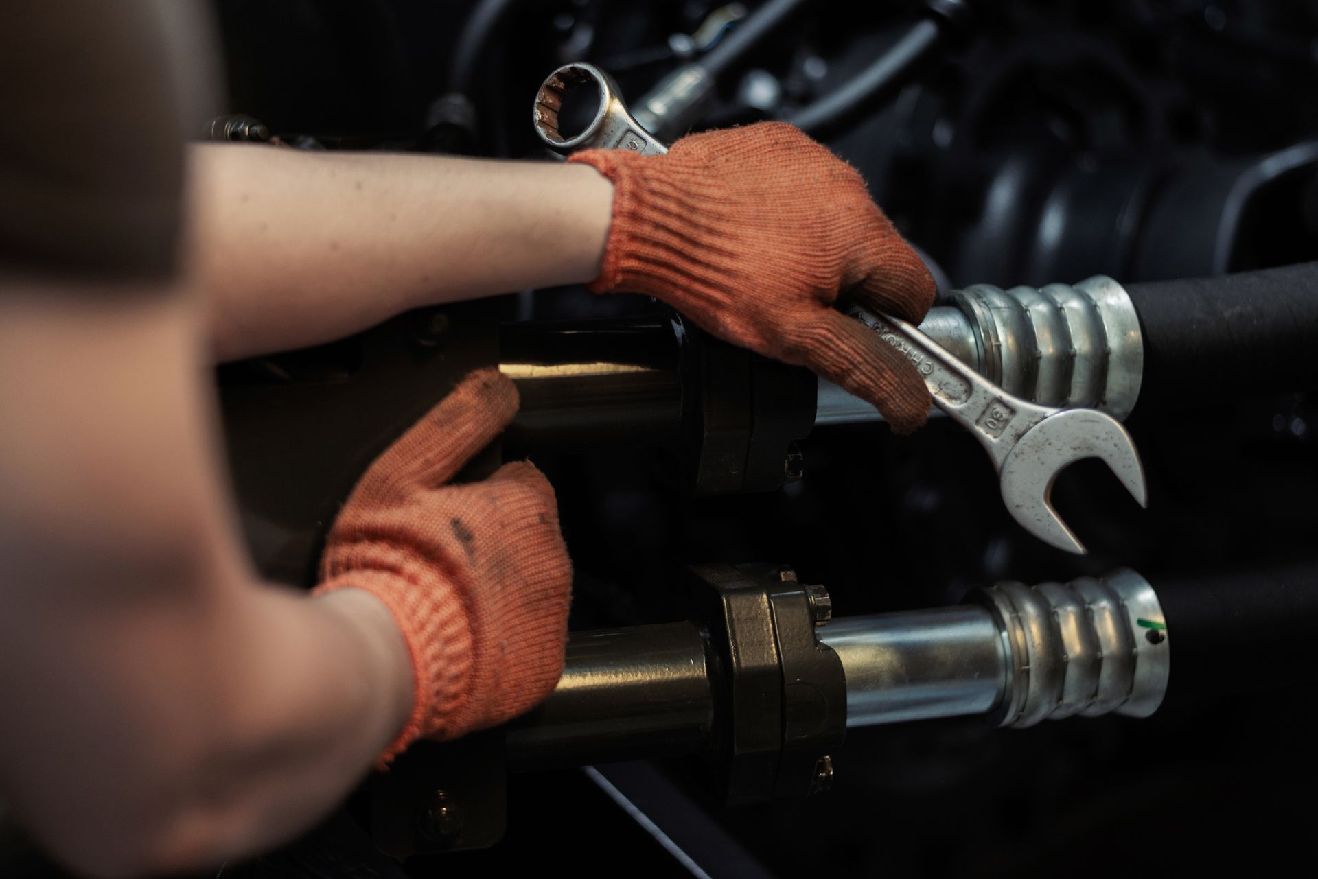 Mechanic's hands in orange gloves using a wrench on a dark industrial component.