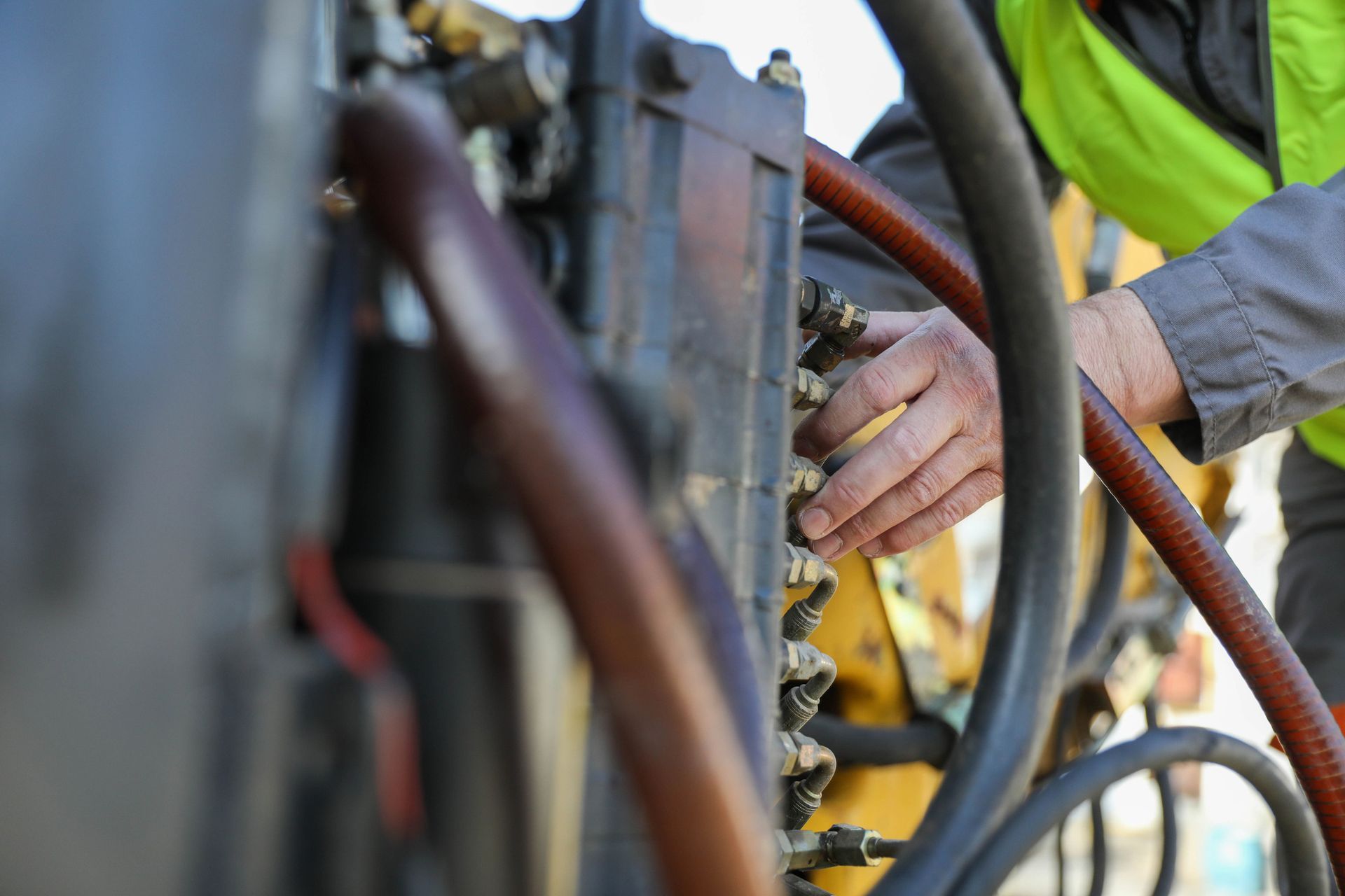 Mechanic's hand examines machinery; focus on a series of connected metal components, cables, and hydraulic lines; wearing a safety vest.