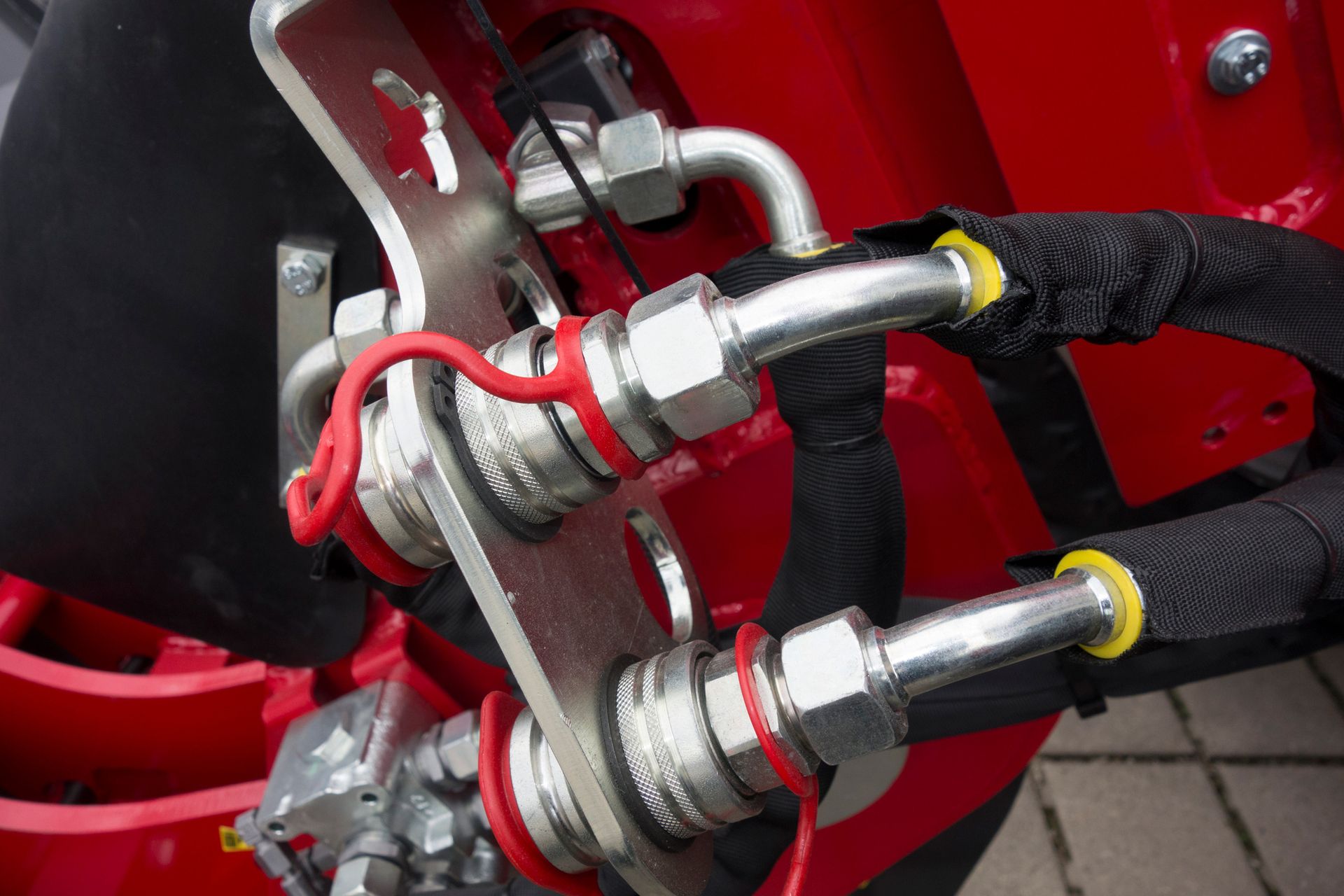Close-up of hydraulic fittings on a red machine. Steel tubes with silver connectors, red seals, and black protective sleeves.