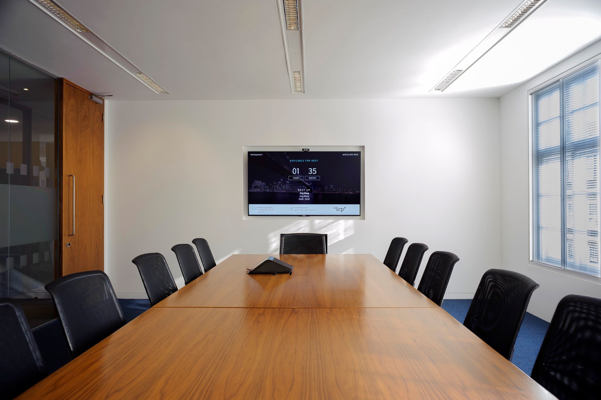 Empty conference room with a long wooden table, black chairs, wall-mounted TV, and large windows.