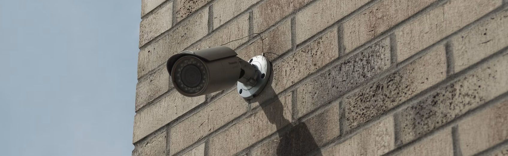 Wall-mounted security camera on a brick building, casting a shadow in sunlight