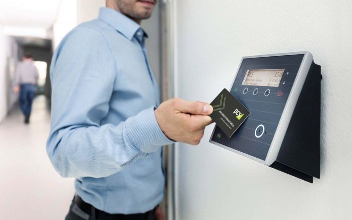 Man in a blue shirt holding a card to a wall-mounted access control panel indoors