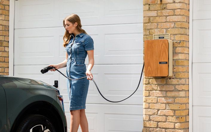 Woman charging an electric car from a wall-mounted charger beside a brick garage