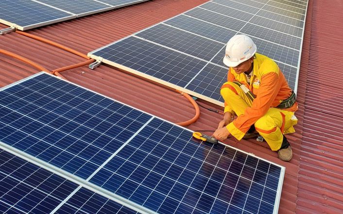 Worker inspecting rooftop solar panels with a tool, wearing orange safety gear and hard hat