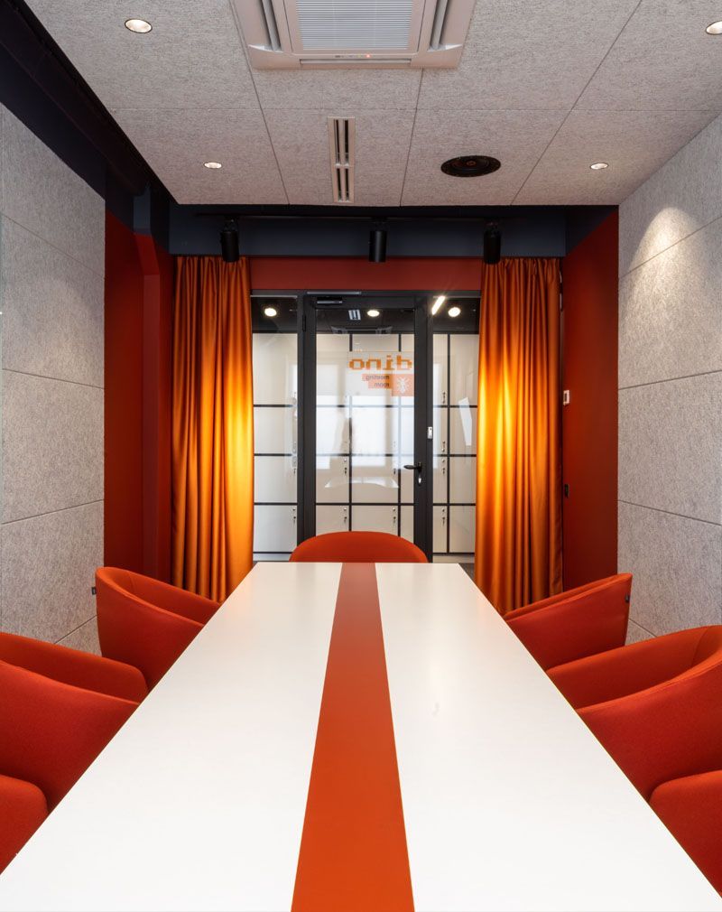 Modern conference room with a white table, orange chairs, and orange curtains by a glass door.