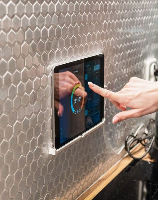 Hands using a touchscreen control panel mounted on a hex-tile wall.