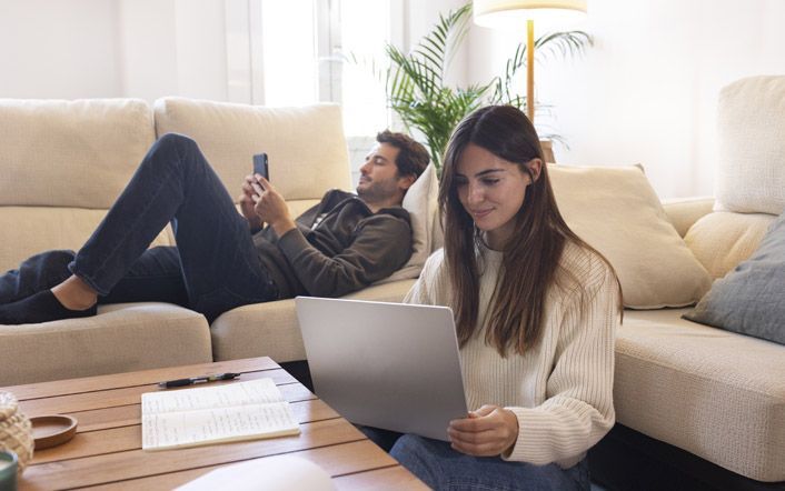 Two people relaxing on a beige couch, one using a laptop and the other looking at a phone.