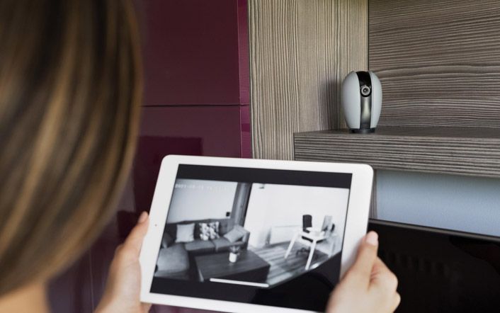Person holding a tablet showing a black-and-white room layout next to a bed and wall lamp