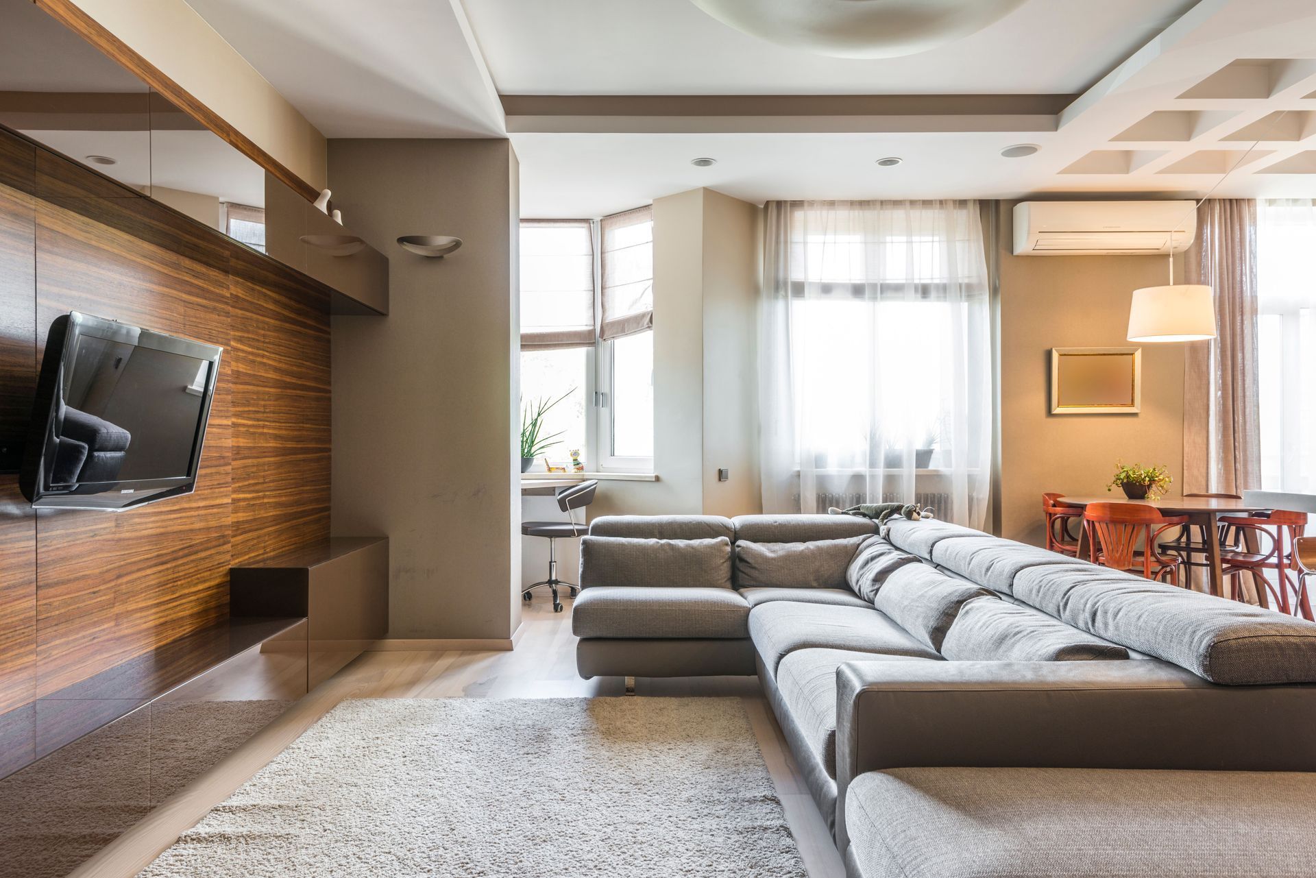 Modern living room with gray sectional sofa, wall-mounted TV, and large windows with sheer curtains