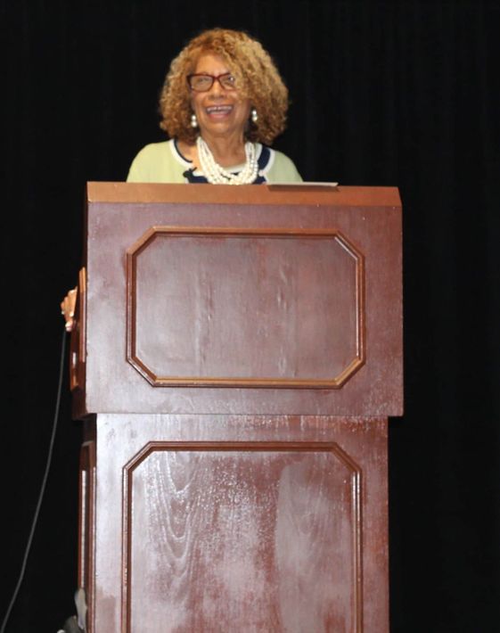 Woman speaking at a wooden podium, smiling. Dark background, wearing glasses, white necklace, light green top.
