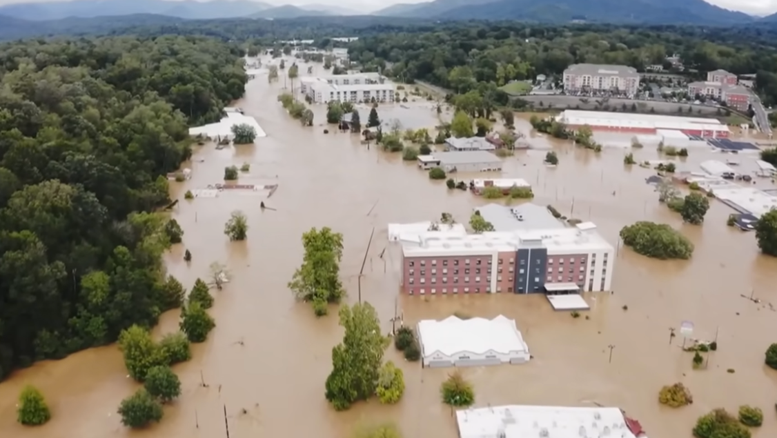 Flooded town with buildings partially submerged in muddy water, trees visible. Overcast sky.