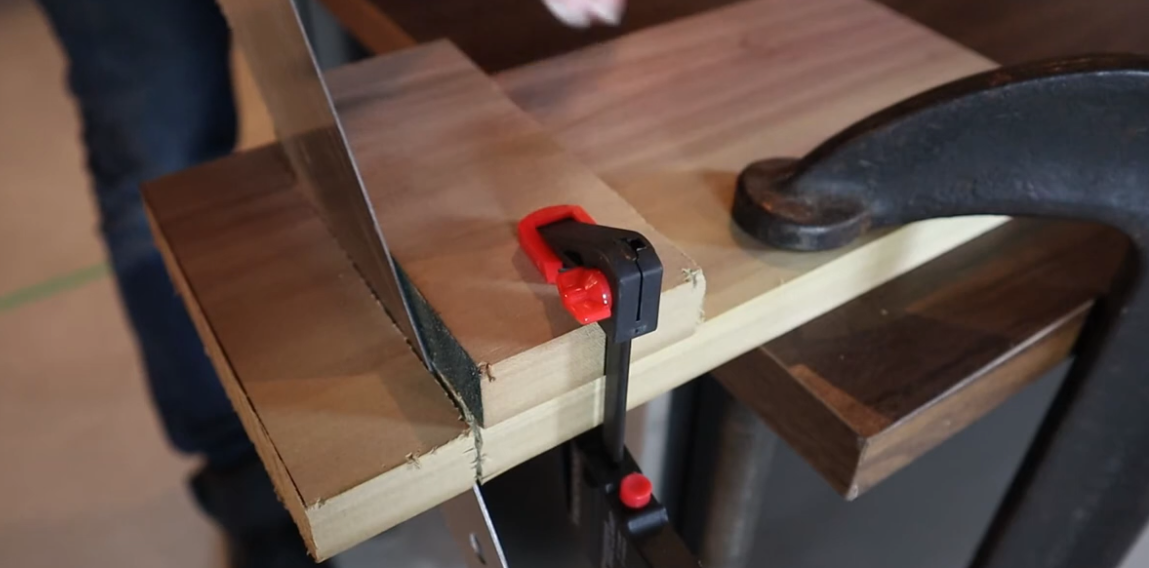 A person sawing a piece of wood clamped to a workbench. A red and black clamp holds the wood in place.