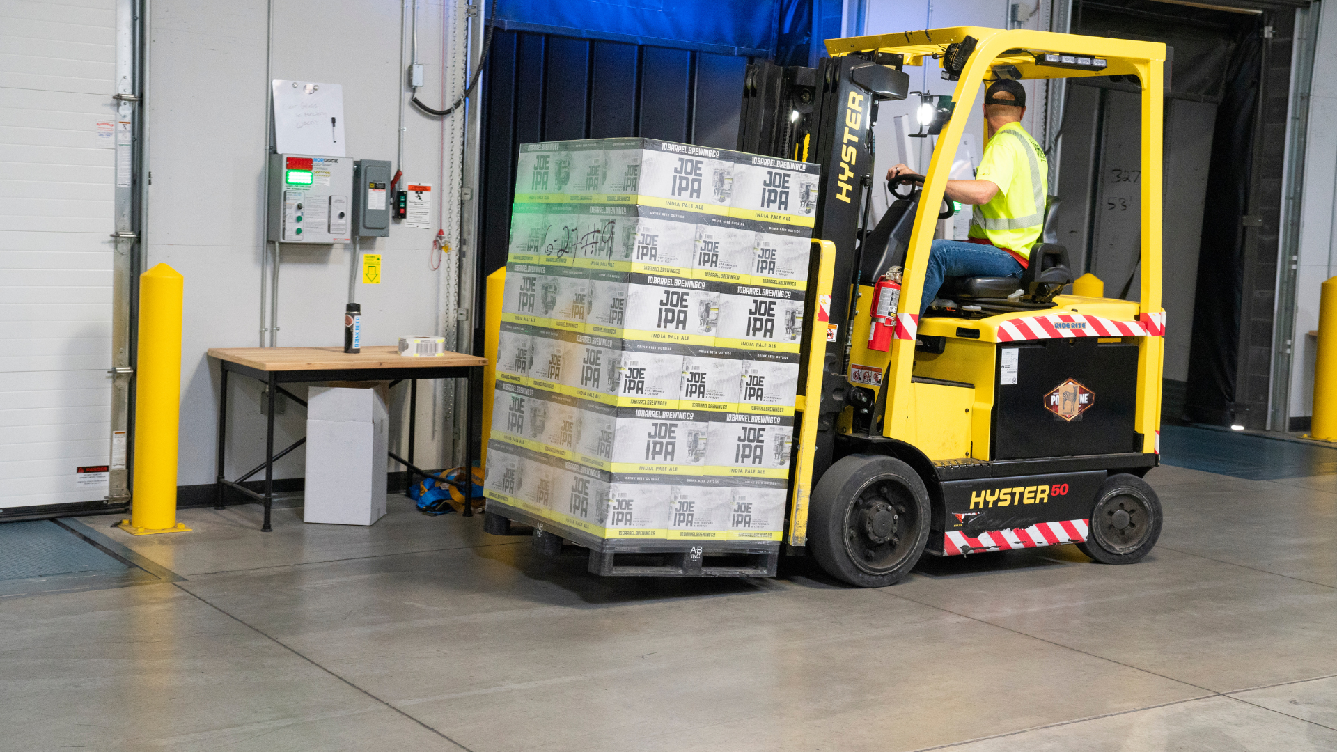 Forklift moving stacked boxes in a warehouse loading area near a desk and wall controls.