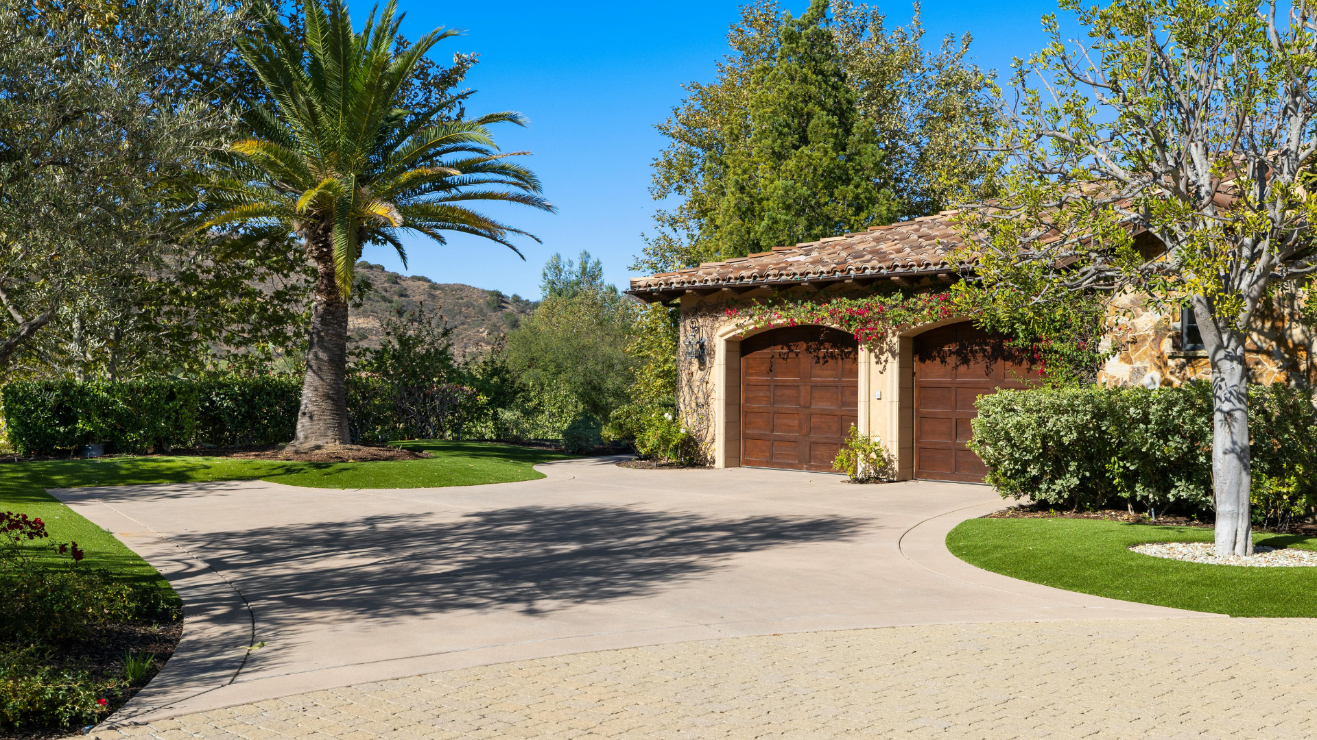 Driveway leading to a garage with two arched wooden doors, surrounded by greenery and trees.