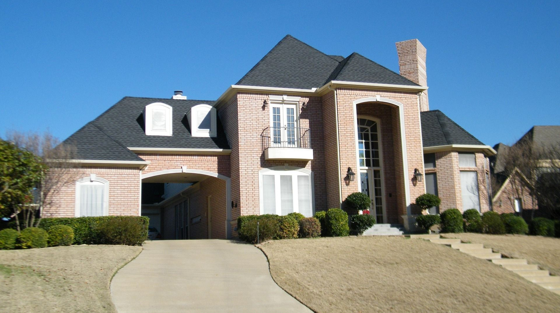 Brick house with dark roof, arched entryway, and a long driveway under a clear blue sky.