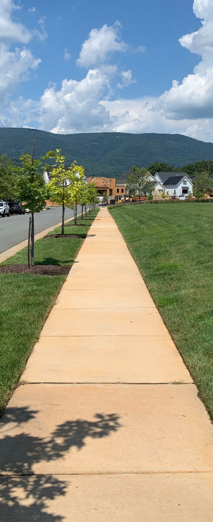 Concrete path through a green park toward houses and mountains under a blue sky with clouds